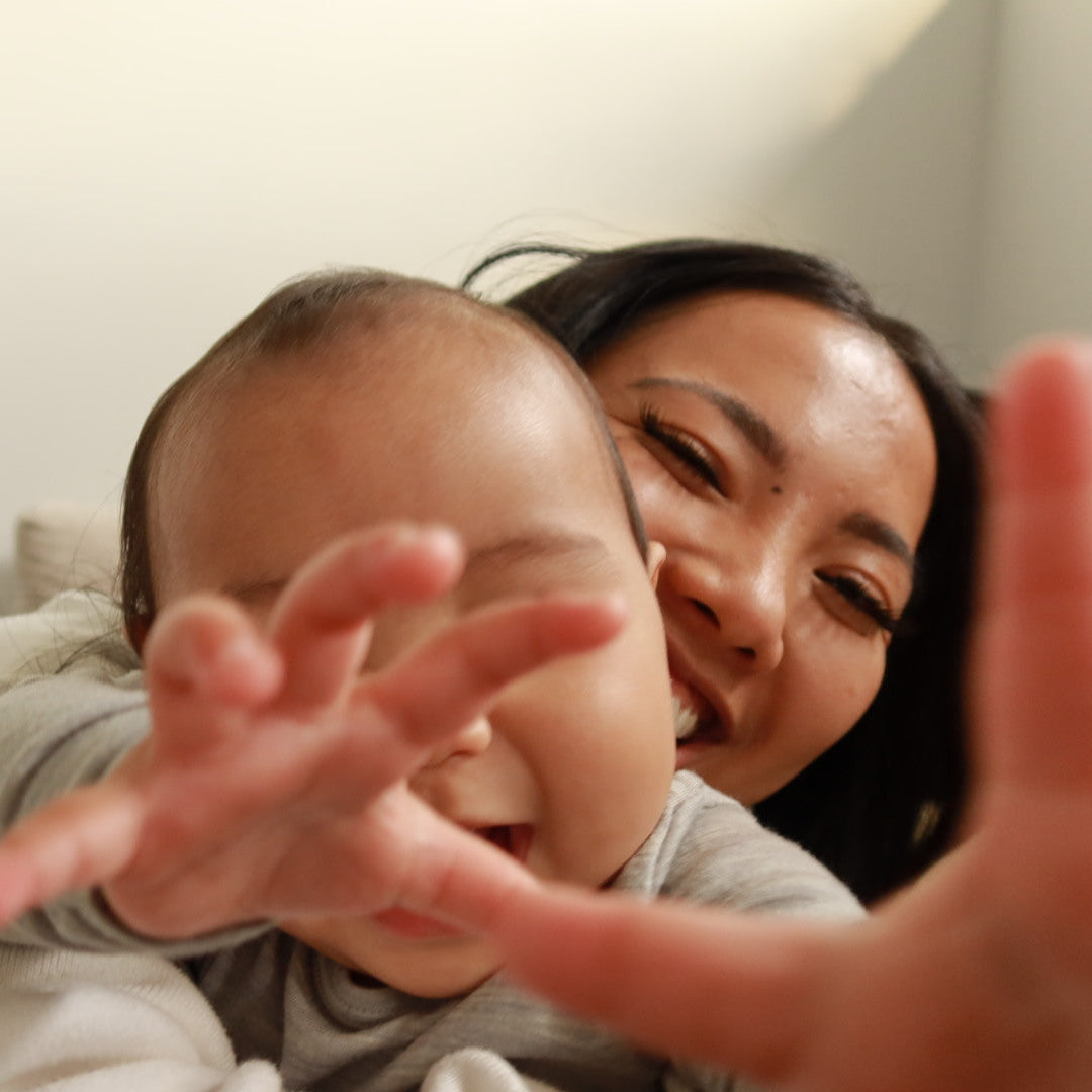 A close-up lifestyle shot of a smiling woman holding a baby who is reaching out with both hands toward the camera in a brightly lit indoor setting.